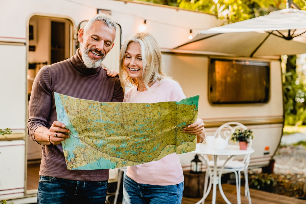 Energetic elderly european couple looking at the map with their caravan home behind. Travelers adventurers explorers reading the map for direction route destination